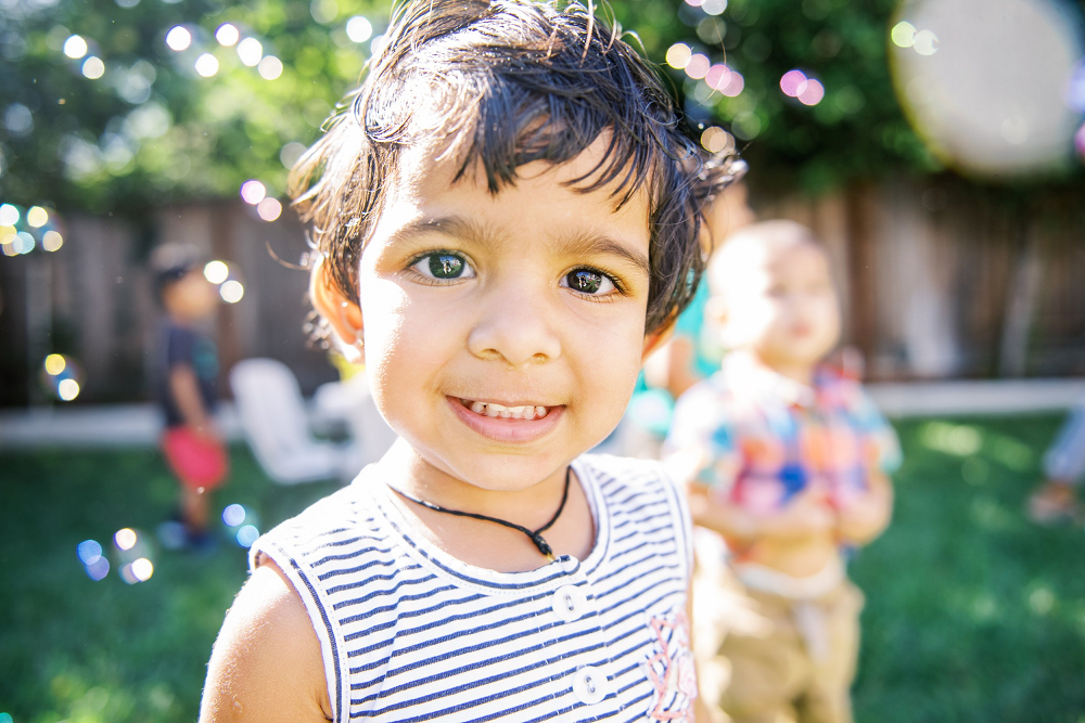 A child in a striped shirt smiles. Bubbles float around them. Another child partially visible in the background.