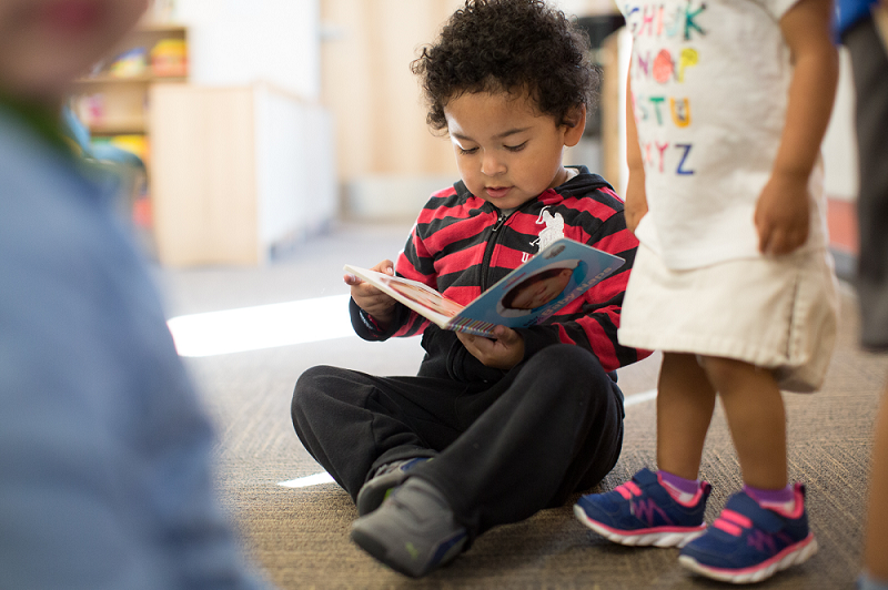 A child sitting on the floor reading a book. They are surrounded by other young children in a bright indoor setting.