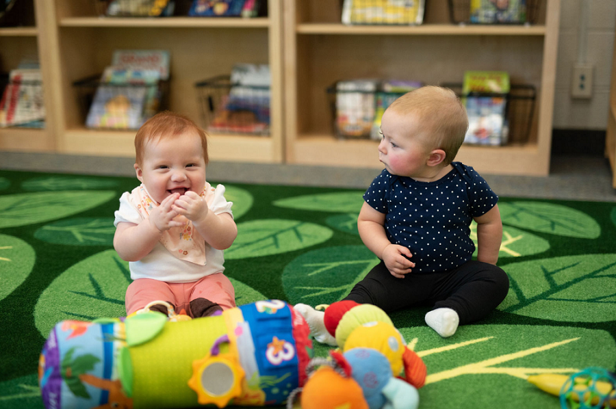 Two babies sitting on a colorful play mat surrounded by soft toys. One baby claps their hands and the other watches intently.